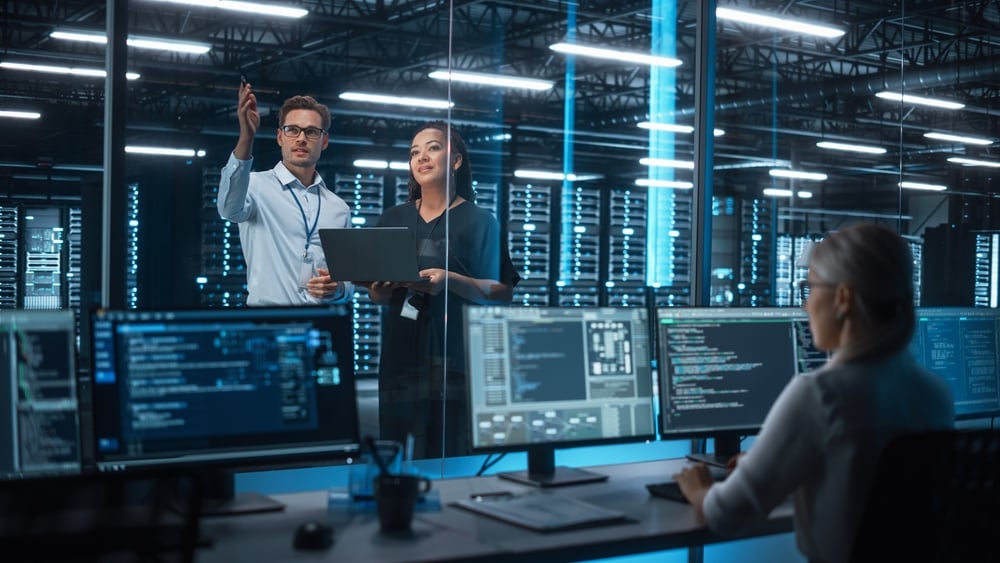 FemaleE-businessEntrepreneurAndMaleItSpecialistDiscussingSomethingAnd Two IT professionals discuss in a server room; man points at a screen while a woman holds a laptop, with colleagues at computer terminals in the background.