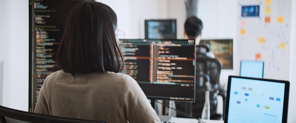 YoungAdultAsianMaleFemaleSoftwareDeveloperCodingProgramOn Person with shoulder-length hair sits at a desk coding on multiple monitors in a bright office setting