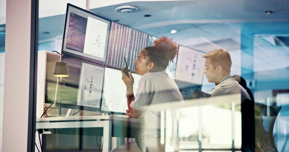 PeopleMeteorologyAndTeamWithScreenForecastAndRadioFor Two coworkers monitor multiple screens displaying code and diagrams in a glass-walled office.