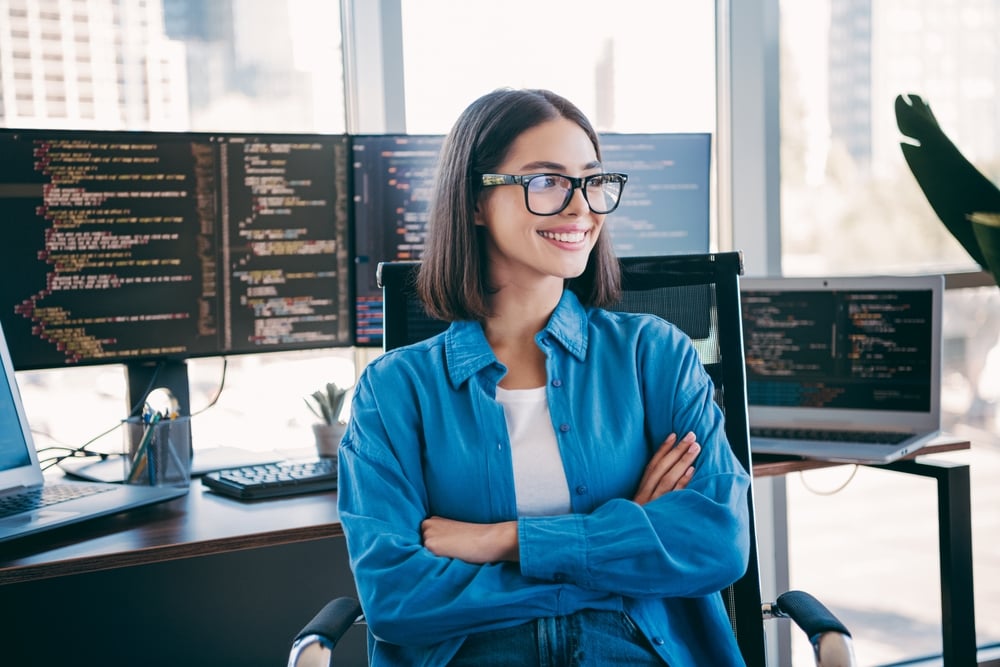 ACheerfulFemaleProgrammerSitsAtABusyDeskSurrounded female programmer sits at a busy desk surrounded by multiple computer monitors