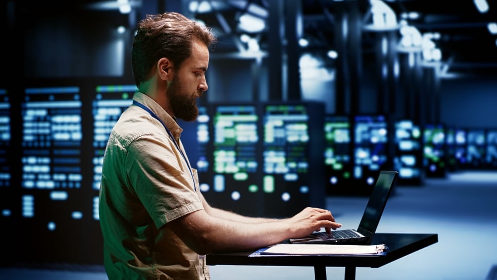 ComputerScientistTypingCloudComputingServiceBusinessDisasterRecoveryPlan Bearded man in a light shirt working on a laptop at a standing table in a server room with blinking racks behind him