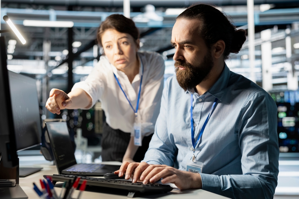 ProgrammersInDataCenterWorkplaceUsingComputerVerifyingFirewallConfigurations Two IT professionals in a server room work at a desk: one man types on a keyboard while a colleague points at a monitor, discussing the screen content.