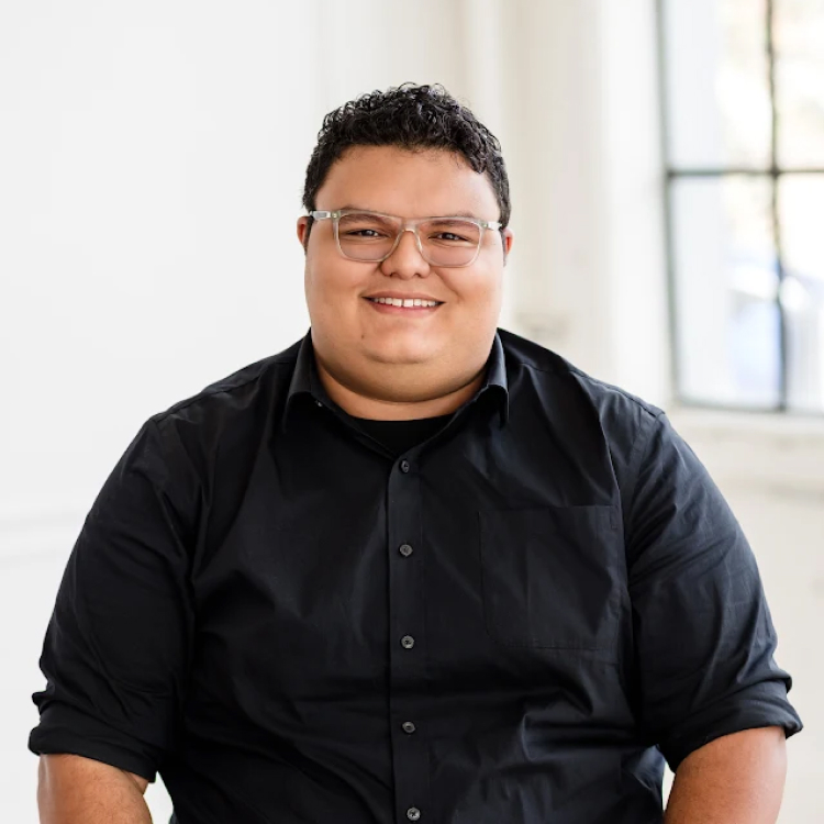 Smiling Person with Short Dark Hair and Clear framed Glasses in a Black Button up Shirt Indoors by a Bright Window