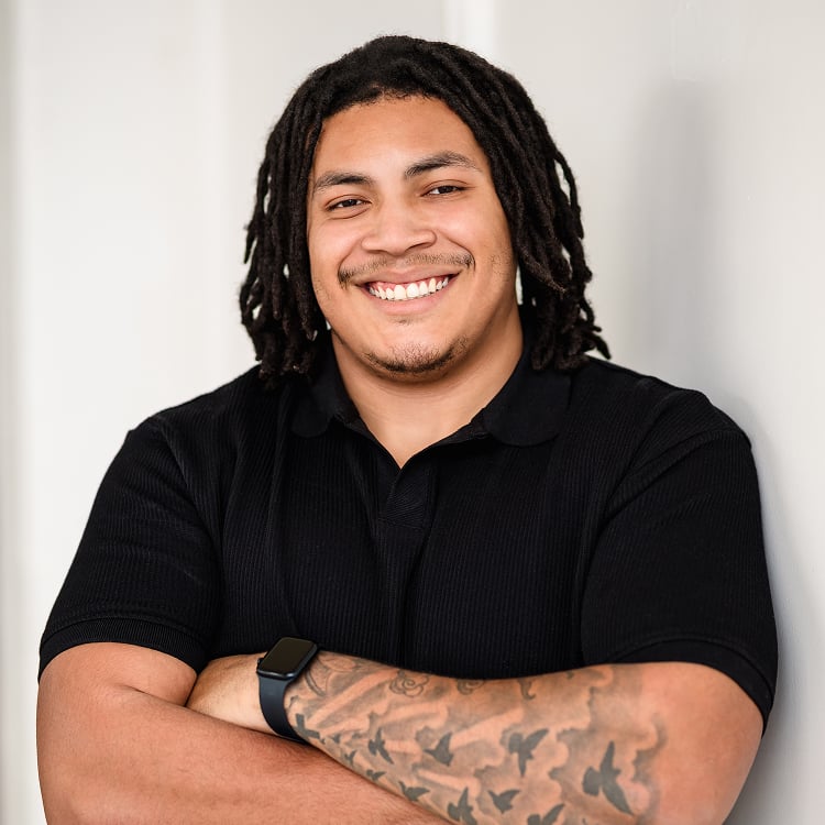 Smiling Man with Dreadlocks in a Black Polo Arms Crossed Standing Against a Light Wall with a Tattooed Forearm and Smartwatch Visible