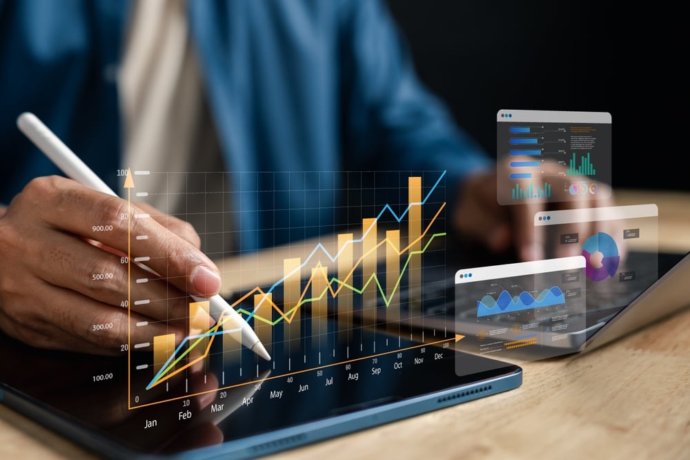 Person using a stylus on a tablet with layered charts and dashboards (line and bar graphs).