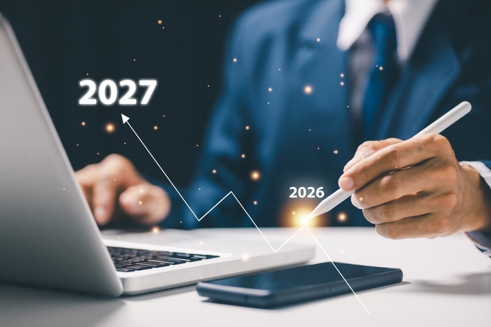 BusinessFuturePlanningConceptWithTimelineGrowthFrom2026To Businessperson in a suit writes with a stylus as a rising projection line shows 2026–2027 beside a laptop and phone.